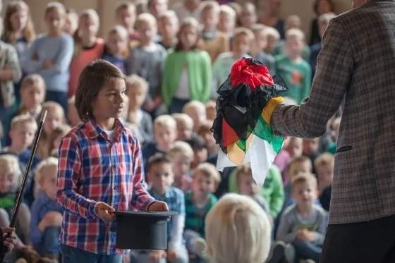 Een jongen helpt mee met een goocheltruc voor een volle zaal kinderen